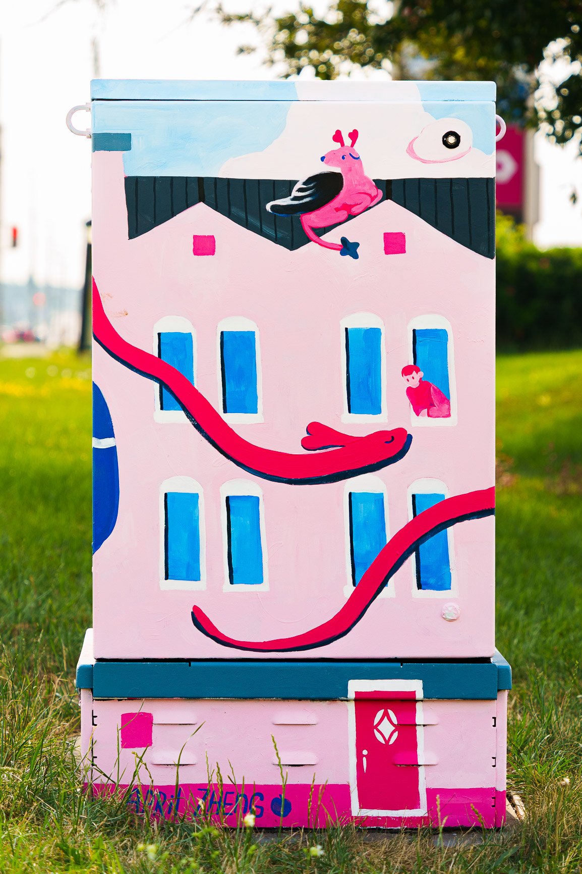 AprilZhengBack A painted utility box features a whimsical scene of a pink building with blue windows. On the roof, a large pink bird with red accents sits beside a smaller blue bird. Two long red snakes slither across the building, one entering through an open window where another small pink bird is perched. At the bottom, a red door with a diamond-shaped window is surrounded by grass. The background includes greenery and blurred outdoor elements.