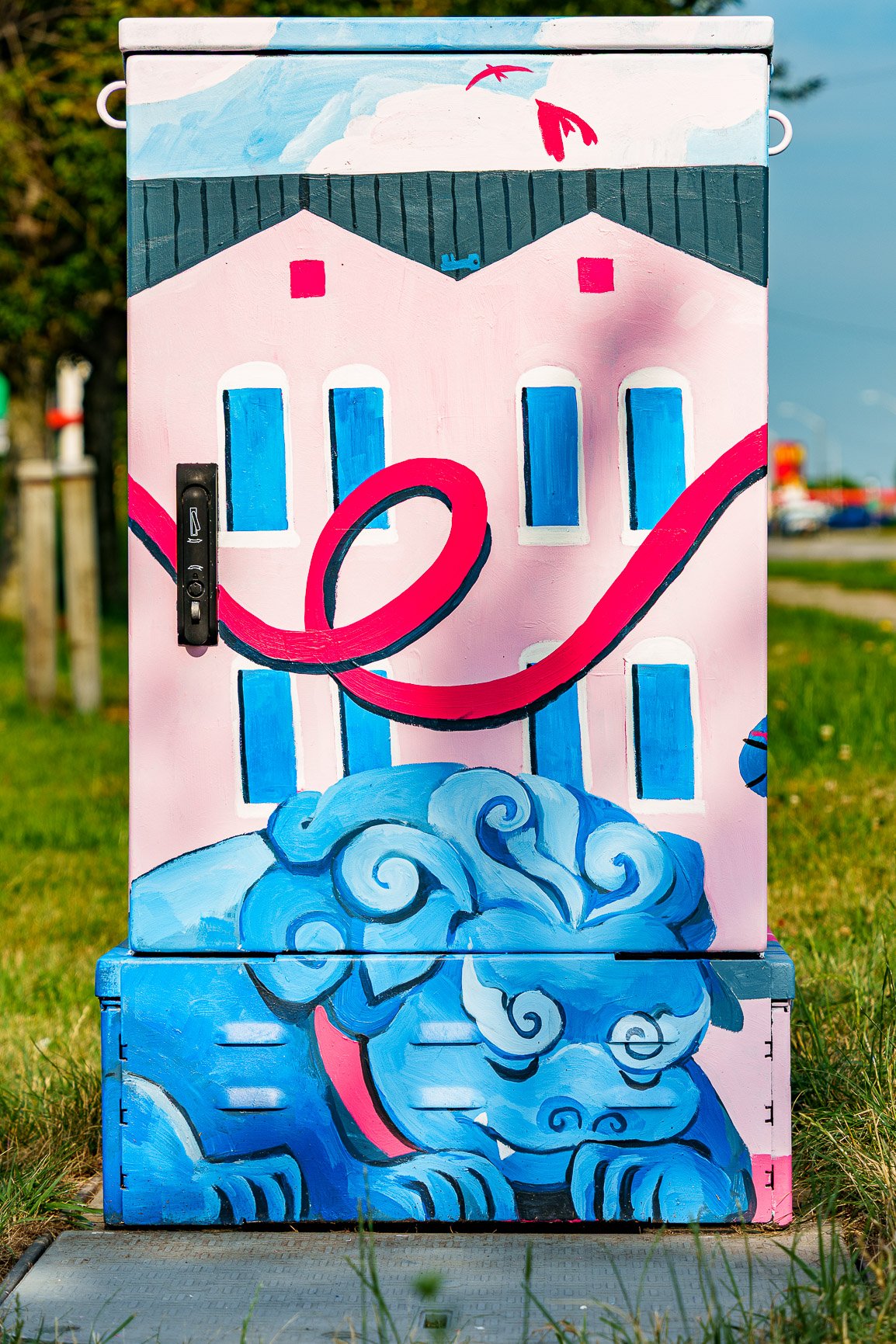 AprilZhengBack A painted utility box displays a vibrant mural. The top half features a stylized pink building with blue windows and a looping red ribbon-like design. The bottom half portrays a blue, cloud-like creature with swirling patterns, reminiscent of lion statues. Green grass and blurred outdoor elements form the background.