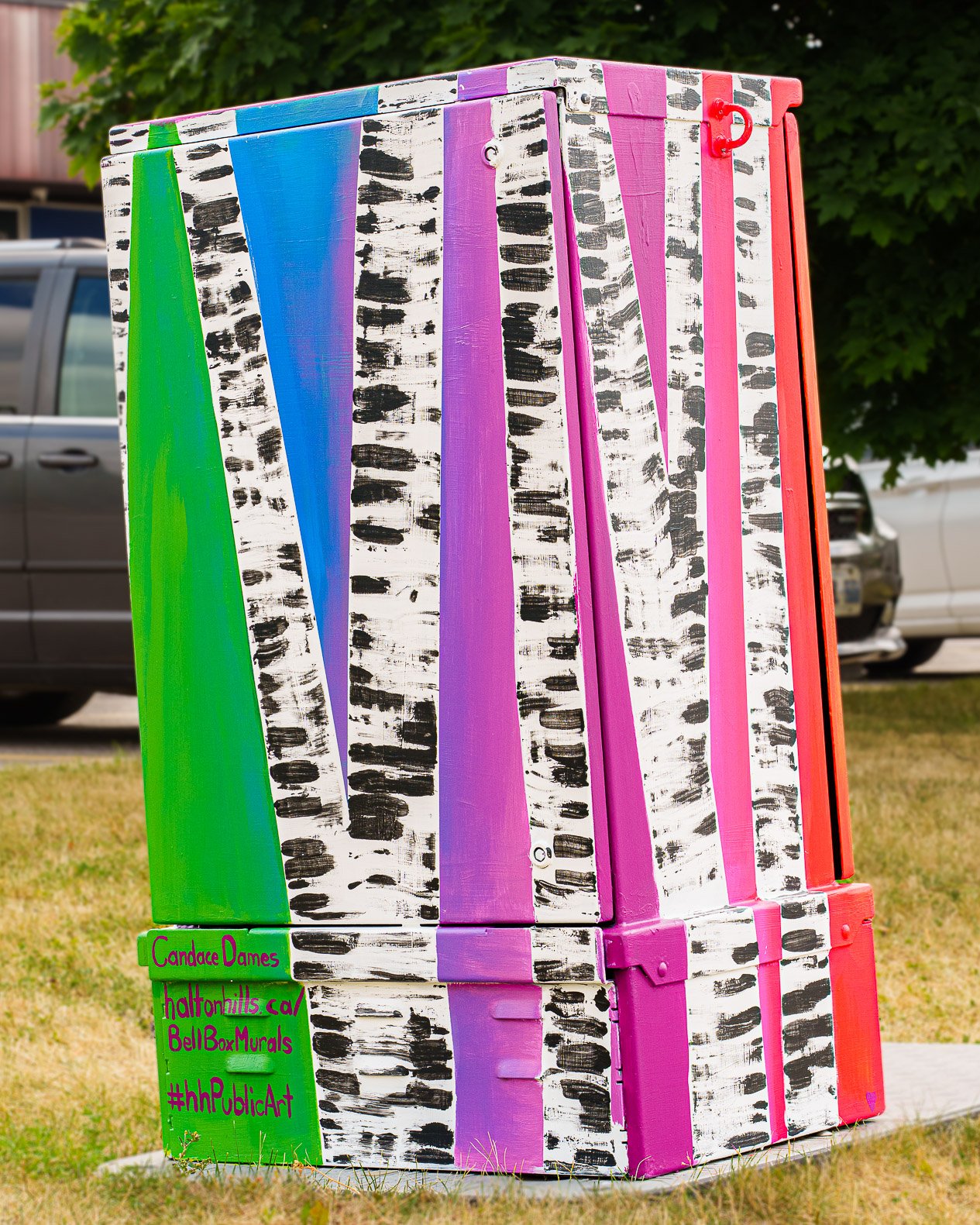 CandaceDamesBellBox A painted utility box features a colorful design with vertical stripes resembling birch trees. The background transitions through shades of green, blue, purple, pink, and red. The artist's name "Candace Danes" and hashtags "#haltonhills.ca", "#BellBoxMurals", and "#hhPublicArt" are visible in the bottom left corner