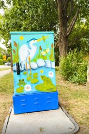 GabeStubbsHeron A painted utility box outdoors features a colorful wetland scene. A white heron stands in water surrounded by cattails, lily pads, and two green frogs. The background includes blue skies and lush greenery. The box is set in a grassy area with trees, transforming urban infrastructure into vibrant public art.