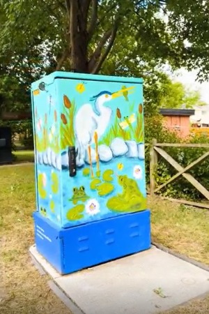 GabeStubbsCorner A painted utility box outdoors features a vibrant wetland scene. A white heron stands in water surrounded by cattails, lily pads, and two green frogs. The background includes blue skies and lush greenery, creating a lively nature-inspired mural. The box is set in a grassy area with trees nearby.